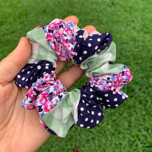 May include: A colorful scrunchie with a mix of patterns. It features sections of navy blue with white polka dots, pink and blue floral prints, and light green gingham. The scrunchie is held in a hand against a green background.