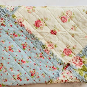 May include: A rectangular quilted zipper pouch featuring a diagonal patchwork design. The fabric combines cream and light blue sections, both decorated with small pink rose floral patterns. A small white tassel is attached to the metal zipper.
