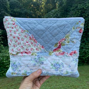 May include: A patchwork quilted pouch with a zipper closure. The pouch features a mix of floral and gingham patterns in shades of blue, pink, and white. The design includes ruffled edges and a quilted texture, creating a vintage aesthetic.