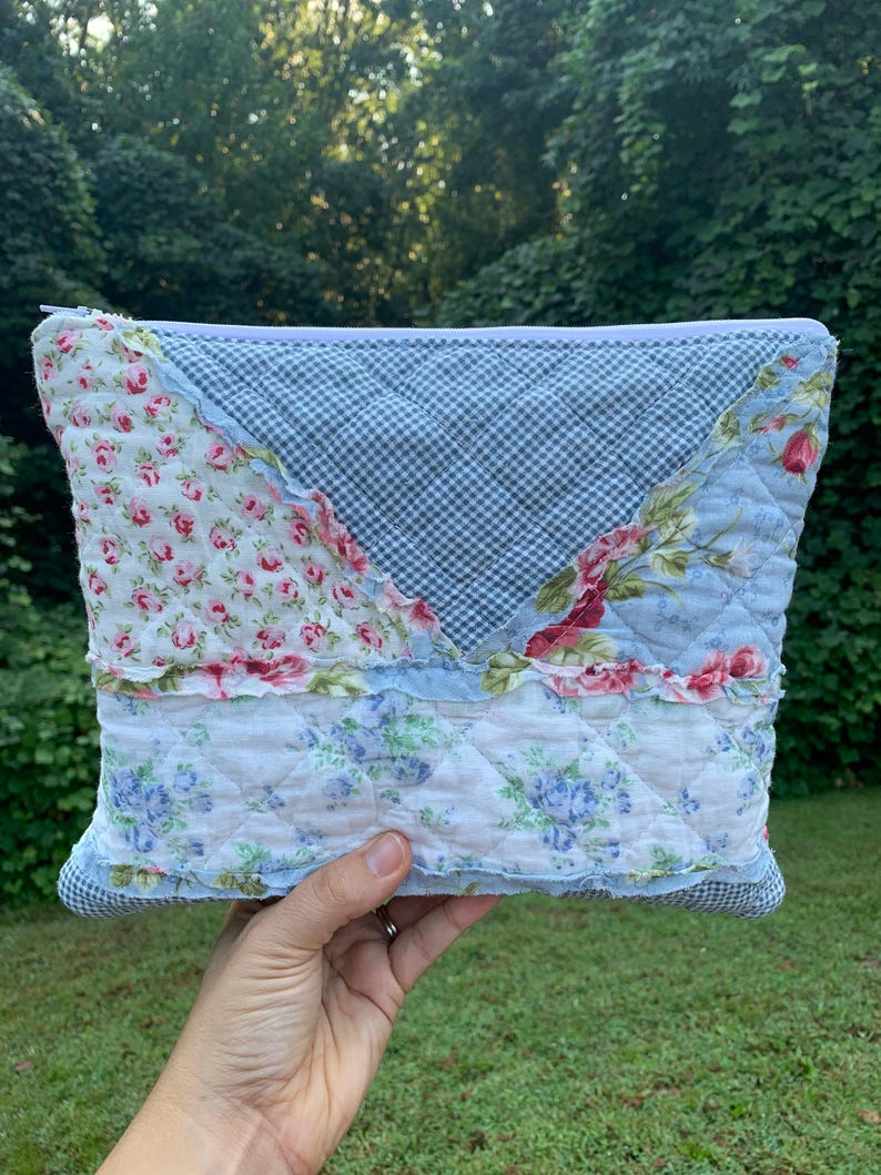 May include: A patchwork quilted pouch with a zipper closure. The pouch features a mix of floral and gingham patterns in shades of blue, pink, and white. The design includes ruffled edges and a quilted texture, creating a vintage aesthetic.