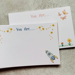 May include: Two white rectangular note cards. The front card features the blue text "You Are...." with yellow stars and a blue and yellow rocket ship. The back card features the pink text "You Are..." with orange butterflies and flowers.
