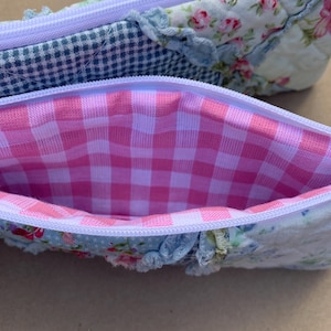 Peut inclure: Deux trousses en tissu. L'une présente un motif vichy bleu et blanc, l'autre un motif floral avec des roses roses. Les deux trousses ont un intérieur vichy rose et blanc et une fermeture éclair lavande.