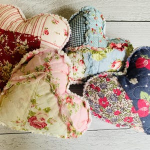 May include: A collection of five heart-shaped fabric ornaments. Each heart features a patchwork design with floral and patterned fabrics in shades of red, pink, blue, and cream. The edges are finished with a ruffled trim.