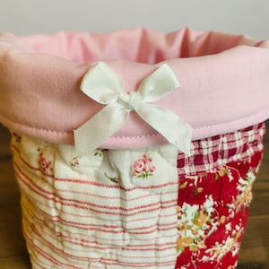 May include: A small, patchwork fabric basket with a pink interior and a white bow. The exterior features a mix of red floral, striped, and checkered patterns. The basket is designed for storage and decorative use.