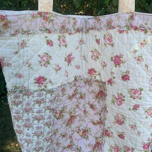 May include: A quilted tote bag with a floral rose pattern in pink and white. The bag features a diamond pattern, ruffled trim, and matching floral handles. The design is reminiscent of vintage quilts.