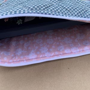 May include: A quilted laptop sleeve with a floral pattern on the exterior and a pink floral lining. The sleeve has a white zipper closure and a blue and white checkered pattern on the top.