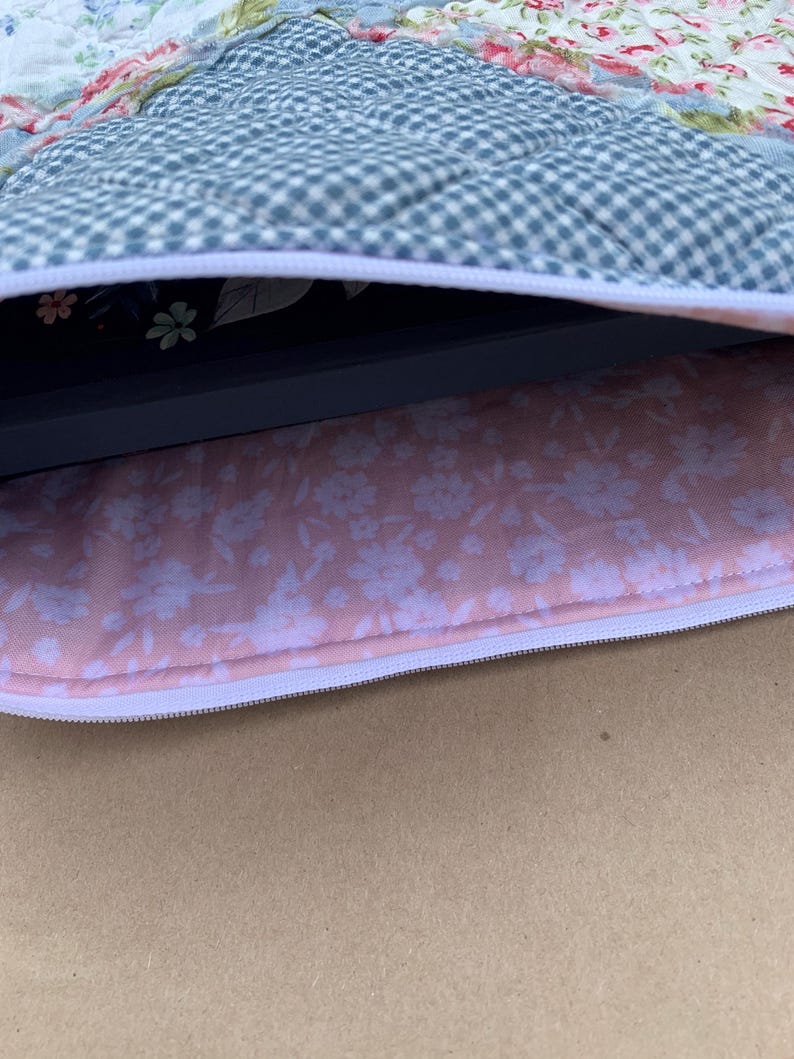 May include: A quilted laptop sleeve with a floral pattern on the exterior and a pink floral lining. The sleeve has a white zipper closure and a blue and white checkered pattern on the top.