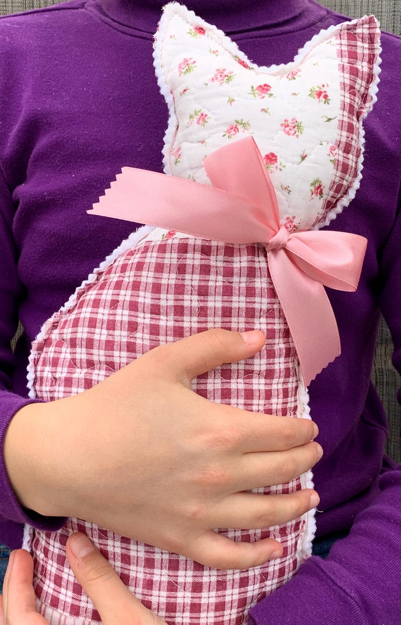 May include: A handmade cat-shaped pillow. The pillow features a white floral print fabric on the head and a red and white checkered pattern on the body. A pink satin bow adorns the neck, adding a decorative touch.