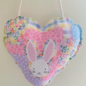 May include: Heart-shaped fabric wall hanging with a bunny face. The patchwork design features floral, polka dot, and gingham patterns in pastel colors. A white rope is attached for hanging. The bunny has pink ears and a smiling face.