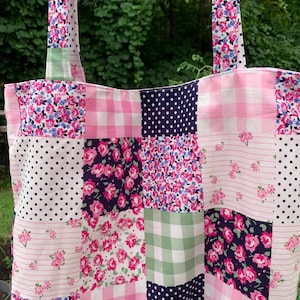 May include: A patchwork tote bag with a variety of floral, polka dot, and gingham patterns in pink, white, and navy blue. The bag has long straps and is made of fabric. The bag is designed for carrying items.