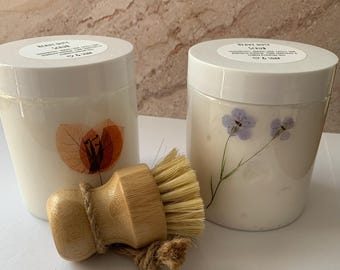 May include: Two white jars with lids, one decorated with a leaf design and the other with purple flowers. A wooden brush with natural bristles and a jute rope handle sits in front of the jars. The jars have labels that read "Heavy Duty Scrub".
