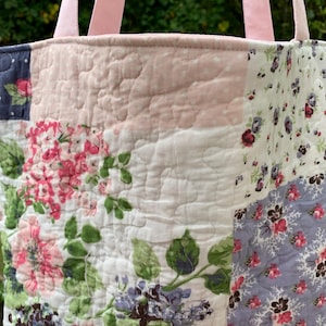 May include: Quilted tote bag with a patchwork design. The bag features various floral patterns in pink, white, and blue, with pink handles. The bag is made of quilted fabric.