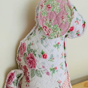 May include: A quilted dog-shaped pillow with a floral pattern. The pillow features a white background with pink and red roses, green leaves, and blue accents. The dog's ears are a solid pink with a floral design. The edges are trimmed with a white, textured fabric.
