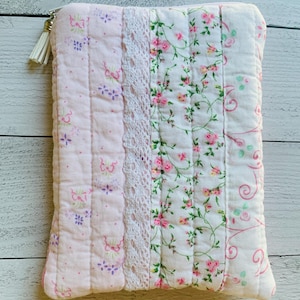May include: A rectangular quilted pouch featuring vertical panels of light pink fabric with small floral patterns, a strip of white lace trim, and a white floral print on a white background. The pouch includes a white zipper with a white tassel pull.