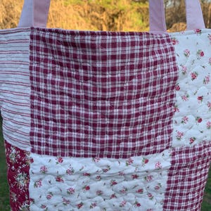 May include: A quilted tote bag with a patchwork design. The bag features a combination of fabrics, including red and white gingham, floral prints, and striped fabric. The handles are a solid pink color.