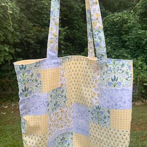 May include: A patchwork tote bag with floral and gingham patterns in yellow, blue, and white. The bag features long straps with a floral design and is set against a backdrop of green foliage.