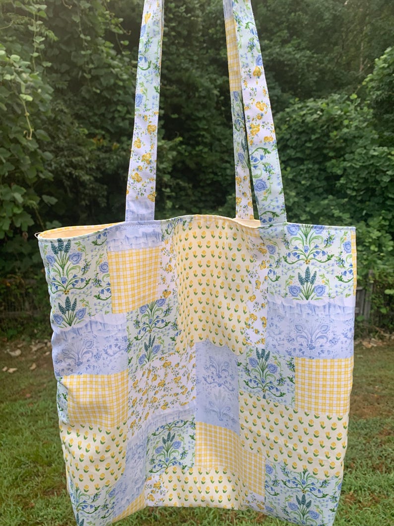May include: A patchwork tote bag with floral and gingham patterns in yellow, blue, and white. The bag features long straps with a floral design and is set against a backdrop of green foliage.