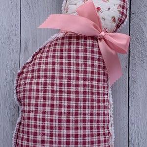 May include: A handmade cat-shaped pillow with a pink bow. The pillow features a red and white gingham pattern on the body and a floral print on the head. The edges are trimmed with white lace.