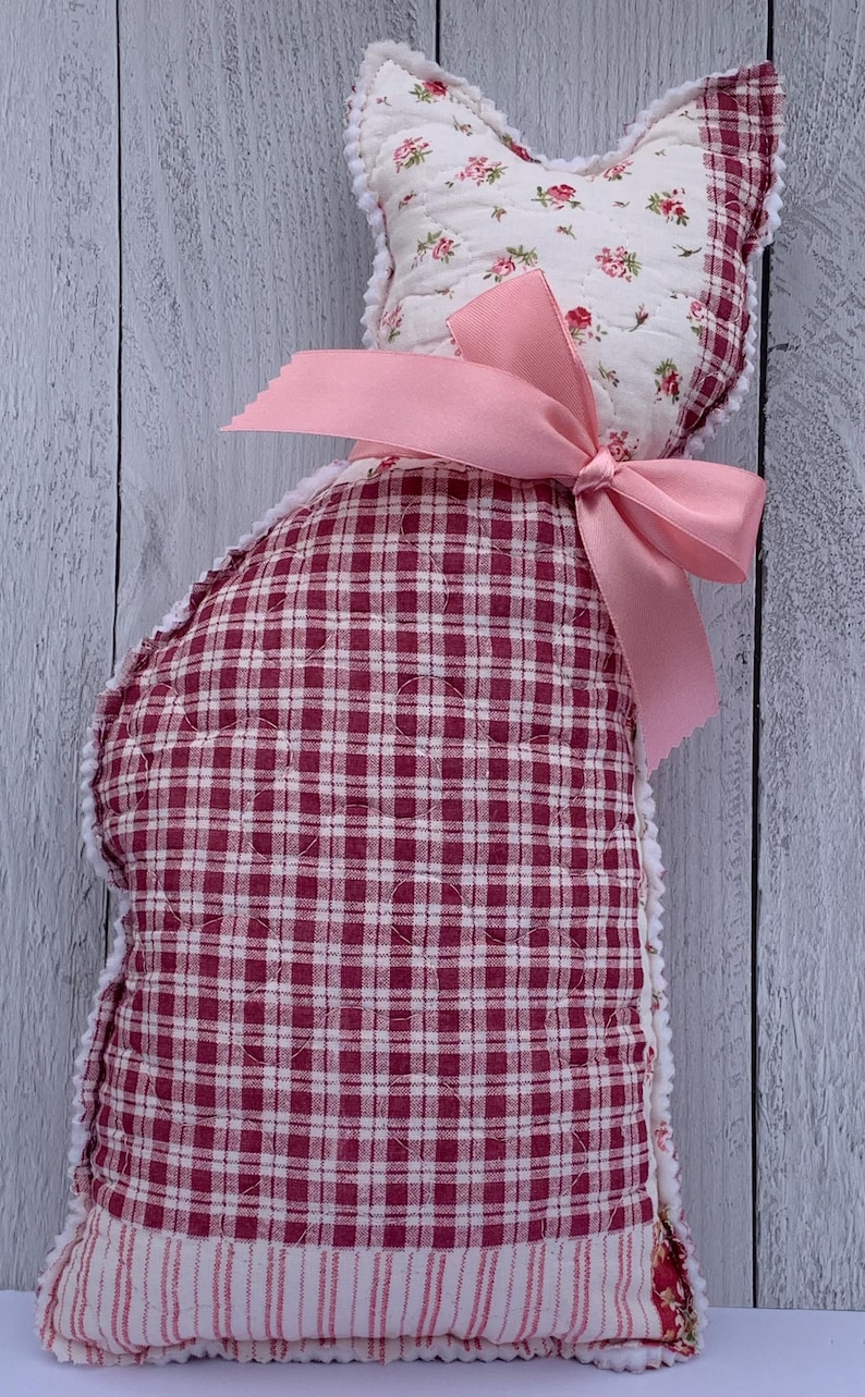 May include: A handmade cat-shaped pillow with a pink bow. The pillow features a red and white gingham pattern on the body and a floral print on the head. The edges are trimmed with white lace.