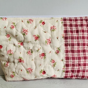 May include: A quilted cosmetic bag with a cream-colored floral pattern on one side and a red and white plaid pattern on the other. The bag has a white zipper and a white tassel. The bag is approximately 8 inches wide.