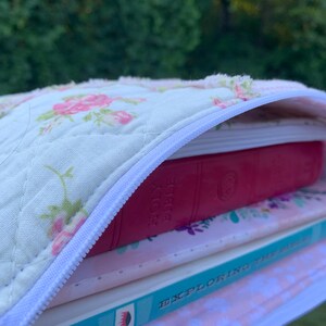 May include: A quilted book cover with a white zipper, filled with books. The cover is cream-colored with a floral pattern in pink and green. A red book labeled "Holy Bible" is visible, along with other books.