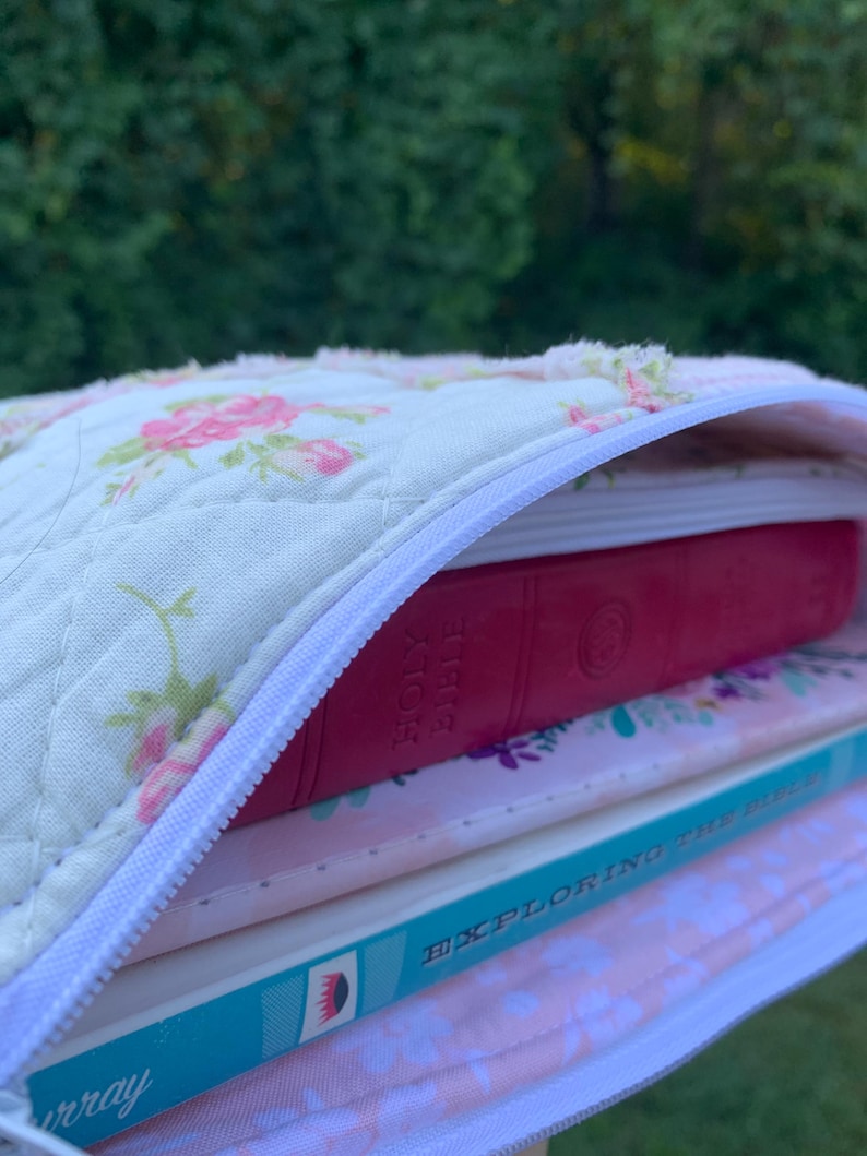 May include: A quilted book cover with a white zipper, filled with books. The cover is cream-colored with a floral pattern in pink and green. A red book labeled "Holy Bible" is visible, along with other books.