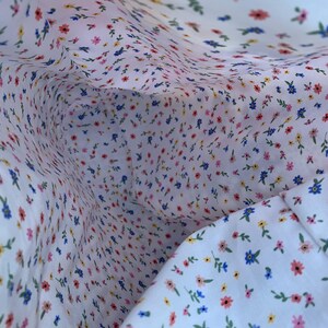 May include: Close-up of white fabric with a delicate floral pattern. The fabric is covered in tiny flowers in shades of pink, blue, and yellow, with green leaves. The pattern is repeated throughout the fabric.
