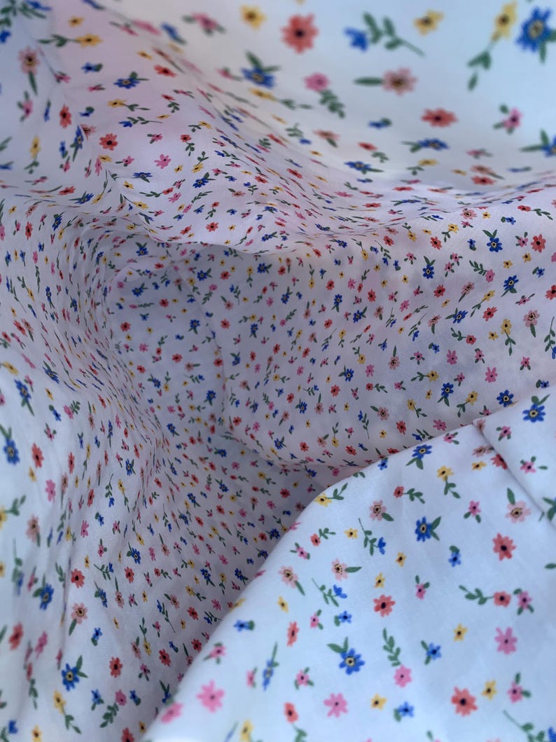 May include: Close-up of white fabric with a delicate floral pattern. The fabric is covered in tiny flowers in shades of pink, blue, and yellow, with green leaves. The pattern is repeated throughout the fabric.