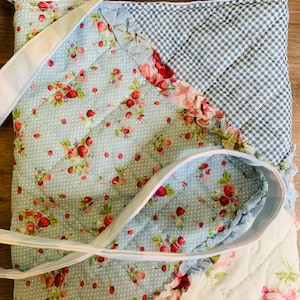 May include: A quilted tote bag with a light blue floral pattern, a gingham pattern, and a white strap. The bag features a patchwork design with red and pink floral accents and ruffled edges.