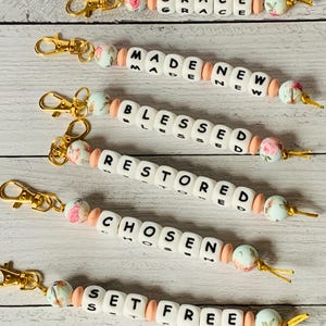 May include: Keychains with inspirational words like "GRACE", "MADE NEW", "BLESSED", "RESTORED", "CHOSEN", and "SET FREE". Each keychain features white letter beads, peach-colored beads, and floral accent beads, with gold-colored clasps.