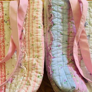 May include: Two quilted tote bags featuring vertical panels of floral and striped fabric in shades of pink, blue, and white. Each bag includes pink fabric handles and decorative white lace trim along the sides with ruffled fabric accents.