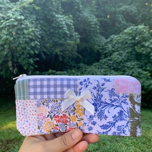 May include: A patchwork pencil case with a white bow. The case features a variety of fabric patterns, including floral, gingham, and toile designs. The case has a zipper closure and is held in a hand.