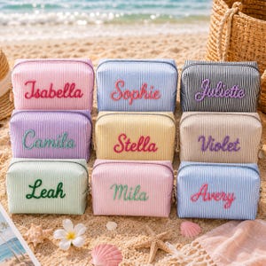 May include: Nine rectangular zippered pouches with vertical pinstripes in various pastel colors. Each pouch features a name embroidered in a cursive font on the front: Isabella, Sophie, Juliette, Camila, Stella, Violet, Leah, Mila, and Avery.