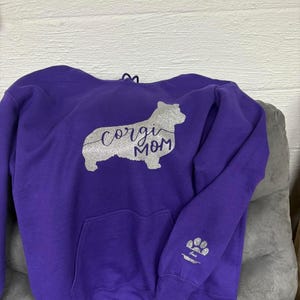 Może przedstawiać: Fioletowa bluza z kapturem ze srebrnym brokatowym wzorem. Wzór przedstawia sylwetkę corgi z napisem "corgi mom" w kursywie. Na rękawie znajduje się wzór w postaci odcisku łapy. Bluza ma przednią kieszeń i kaptur ze sznurkiem.