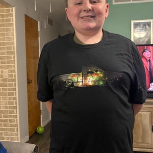 May include: Black t-shirt featuring a graphic of a lowered pickup truck with colorful lights in the bed. The shirt is short-sleeved and has a crew neck. The person wearing the shirt is smiling.