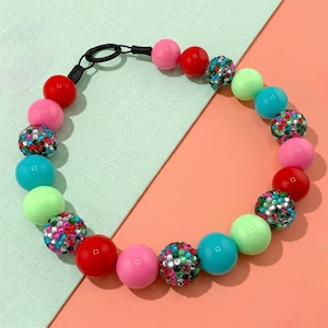 May include: A colorful beaded necklace featuring a variety of round beads in shades of red, pink, green, and turquoise. Some beads are covered in sparkling rhinestones. The necklace has a black clasp.