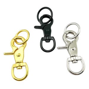 May include: Three swivel snap hooks in gold, black, and silver. Each hook has a spring-loaded clasp and a circular ring for attaching to keychains or other items. The hooks are made of metal and are designed for durability.