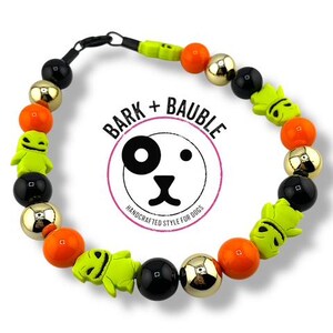 May include: A dog collar featuring a circular design with black, gold, orange, and lime green beads. The collar includes lime green character beads and the text "BARK + BAUBLE" in a circular logo.