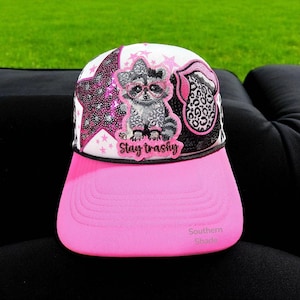 May include: A pink and white trucker hat with a raccoon graphic wearing glasses and a bow. The hat has a pink brim and the text "Stay trashy". The hat has a star and leopard print design.