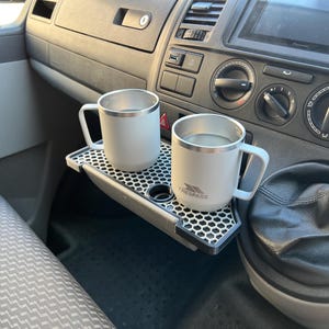 May include: Two white travel mugs with silver lids and handles are sitting on a black and grey tray with a honeycomb pattern. The tray is attached to the dashboard of a car. The mug on the right has the word "Trespass" printed on it.