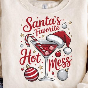 May include: Cream-colored sweatshirt with a festive graphic. The design features a martini glass filled with a red cocktail, a Santa hat, candy cane, and the text "Santa's Favorite Hot Mess" in red script.