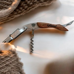 Op de afbeelding: Een zilveren en houten multifunctionele flessenopener met een kurkentrekker, een flessenopener en een klein mes. De handgreep is gegraveerd met het woord "Vincent" en een koksmuts.