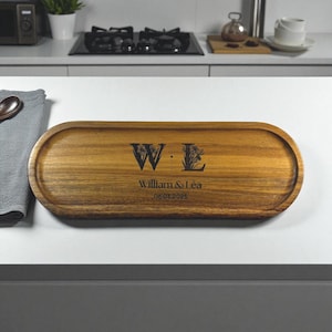 May include: A wooden serving tray with an oval shape. The tray has a dark wood grain and features the initials "W L" with floral accents, along with the names "William & Léa" and the date "06.03.2025" engraved on it.