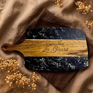 Peut inclure: Une planche de service rectangulaire composée d'une bande centrale en bois naturel clair et de deux sections en marbre noir veiné de blanc. Le centre en bois porte l'inscription gravée L'apéro chez Les Gerard.