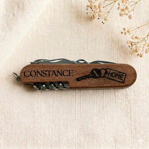 Può includere: Un multiutensile in legno con il nome "Constance" inciso. L'utensile presenta una chiave e la parola "HOME". Include un cavatappi e altri strumenti.
