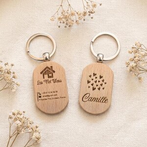 Könnte beinhalten: Zwei Holz-Schlüsselanhänger mit silbernen Ringen. Einer hat ein Haus-Symbol und den Text "Les Flot Bleus" mit Kontaktinformationen. Der andere hat ein Herz-Design und den Namen "Camille".