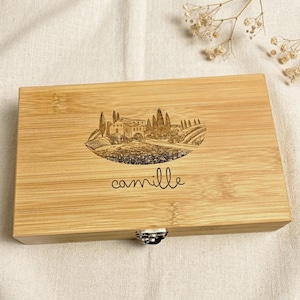 Puede incluir: Una caja rectangular de bambú con una ilustración de paisaje negro y el nombre "camille" en cursiva. La caja tiene un cierre plateado y está hecha de bambú marrón claro.