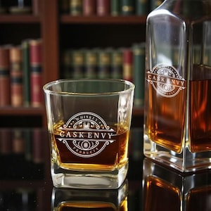May include: A square whiskey glass and decanter filled with amber liquid. Both the glass and decanter have the text "CASK ENVY PRODUCTS" in a white, decorative font. The background features a bookshelf filled with books.