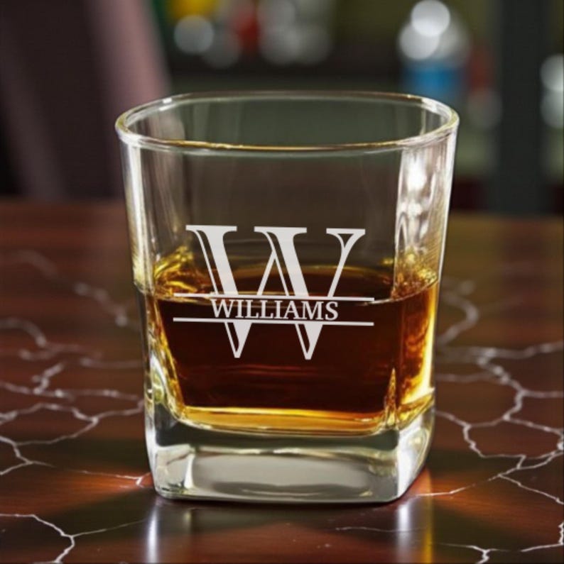 May include: A square-shaped, clear glass tumbler filled with amber liquid. The glass is etched with the name "WILLIAMS" beneath a stylized "W" design. The glass sits on a dark, polished surface.