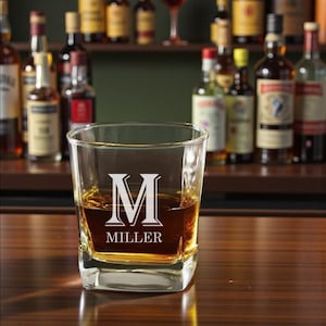 May include: A square-shaped, clear glass tumbler filled with amber liquid, likely whiskey. The glass is etched with a large white letter "M" above the name "MILLER". The background features a bar setting with bottles of liquor on shelves.